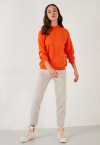 Bright orange sweatshirt with a high collar, long sleeves, and ribbed cuffs, paired with light beige tapered pants and beige sneakers.