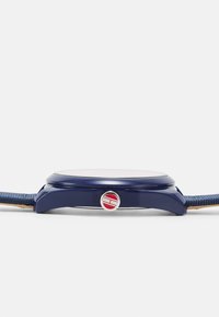 Reloj azul marino con caja redonda, un botón de acento rojo y plateado, y una correa de tela texturizada. Carátula del reloj plana y lisa visible.