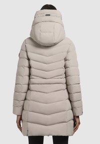 Manteau rembourré beige clair avec un design en matelassage en chevron. Caractéristiques d'une grande capuche, de manches longues et d'un ourlet légèrement courbé. Texture douce.