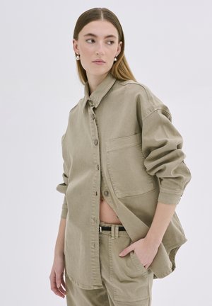 Gaišs khaki oversized krekls ar pogām priekšpusē, vienu krūšu kabatu un sarullētām piedurknēm, izgatavots no teksturēta kokvilnas auduma.