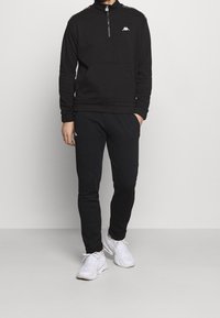 Felpa nera con zip e colletto alto, con logo e dettagli bianchi. Abbinata a pantaloni jogger neri e scarpe sportive bianche.