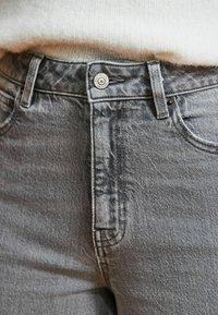 Jean en denim gris avec un design classique à cinq poches, une fermeture à bouton métallique et un tissu texturé visible avec des détails d'effet délavé subtil.
