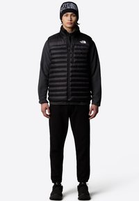 Zwarte puffer vest met horizontale stiksels, hoge kraag en ritsluiting over een zwart langslavenshirt, gecombineerd met zwarte trainingsbroek en schoenen.
