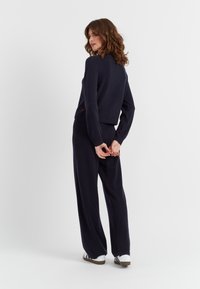 Maglione a maglia navy con orlo a coste e maniche lunghe, abbinato a pantaloni larghi coordinati. Il modello posa con le mani dietro la schiena, indossando sneakers.