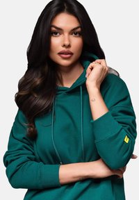Teal hoodie i mjukt material med dragsko i huvan, ribbade ärmslut och en liten gul tagg på ärmen.