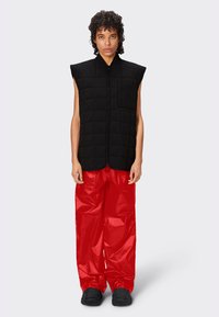 Gilet sans manches matelassé noir avec une fermeture éclair à l'avant et une poche sur la poitrine, associé à un pantalon rouge vif et ample. Des chaussures noires complètent la tenue.