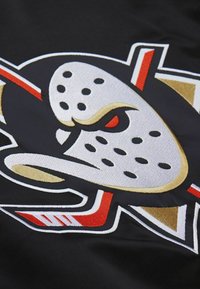 Schwarzer Stoff mit einem auffälligen gestickten Logo, das eine Hockeymaske zeigt, sowie roten und goldenen Akzenten und scharfen, kantigen Designs.