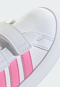 Bílá teniska s hladkou koženou texturou, zdobená růžovými pruhy, zapínáním na suchý zip a kontrastními švy.