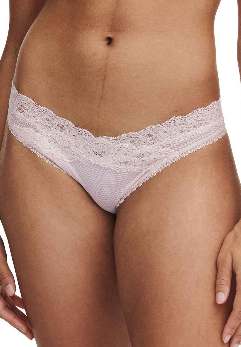 Mujer vistiendo braguitas de bikini de encaje en color rosa pálido con tejido texturizado, mostrada de la cintura a la parte superior de los muslos, con las manos relajadas a los lados.