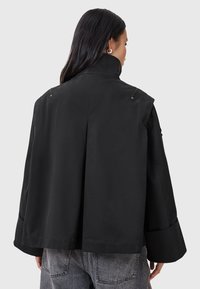 Giacca nera con colletto alto, vestibilità oversize e maniche larghe. Presenta due cerniere di ventilazione sul retro e una texture liscia e opaca.