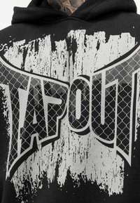 Fekete kapucnis pulóver, amelyen egy feltűnő ezüst és fehér "TAPOUT" grafika található, megkopott textúrával és hálós mintával kiegészítve.