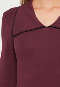 Top in magenta a maglia con un ampio colletto bordato di nero. Presenta maniche lunghe e un design aderente, evidenziando una texture liscia.