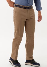Chino beige con una vestibilità slim e dettagli cuciti, abbinati a una cintura scura e sneakers blu navy con accenti verdi e suole testurizzate.