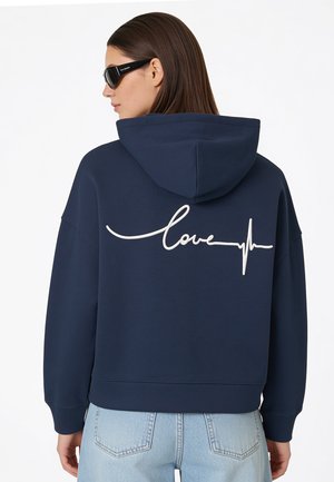 Vrouw draagt een marineblauwe hoodie met wit "love" en een hartslijnontwerp op de rug, lichtblauwe jeans en zwarte zonnebril.