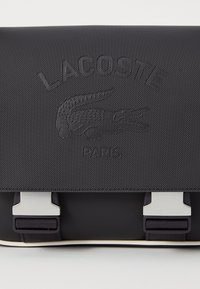 Black Lacoste bag made of durable textured material. Features embossed logo, silver hardware accents, and a flap with secure closures.