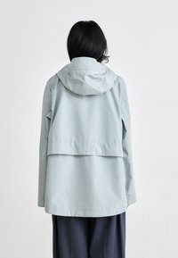 Light blue waterproof jacket with a hood, featuring a relaxed fit, back flap, and long sleeves, paired with dark trousers.