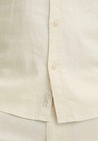 Chemise en lin beige clair avec un devant à boutons, présentant des boutons beiges, un tissu texturé et une étiquette à l'ourlet inférieur.