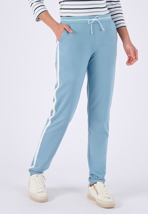 Personne portant un pantalon de jogging bleu clair avec des bandes blanches sur les côtés, un haut à manches longues rayé et des baskets blanches, debout avec une main dans la poche.