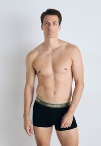 Boxers negros con una cinturilla ancha de color gris claro que presenta el logo "LACOSTE". El modelo tiene el cabello corto y una expresión neutra.