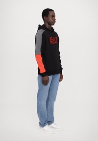 Felpa nera con maniche grigie e arancioni, dotata di un logo sul petto. Abbinata a jeans azzurri chiari e sneakers bianche.