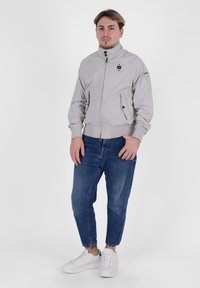 Giacca bomber grigia chiara con colletto alto, chiusura con zip e tasche con zip, indossata sopra pantaloni jeans blu e sneakers bianche. Texture liscia.