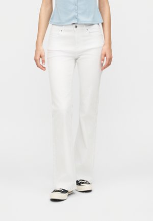 Fehér egyenes szárú farmert, világoskék ujjatlan felsőt és fekete-fehér tornacipőt viselő nő áll sima háttér előtt.