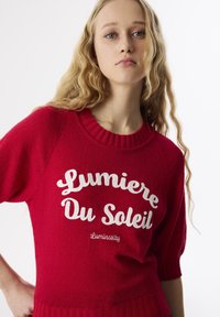 Czerwony sweter dzianinowy z prążkowanym dekoltem okrągłym. Posiada biały tekst "Lumiere Du Soleil" oraz "Luminosity" w kursywie i czcionkach blokowych.