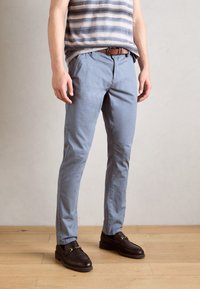 Hellblaue Baumwollhose mit schmaler Passform, kombiniert mit braunen Loafers. Das Modell trägt ein gestreiftes Tanktop und einen braunen Gürtel.