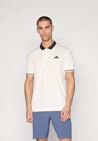 Világos bézs színű pólóing fekete gallérral és három gombos felső résszel. Fekete csíkok találhatók az ujjak szegélyén, valamint egy Adidas logó. Kék sorttal viselik.