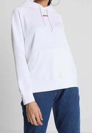 Personne portant un sweat à capuche blanc avec poche avant et un petit logo rouge, associé à un jean bleu sur un fond clair uni.