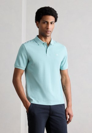 Νεαρός με κοντά σγουρά μαλλιά, που φοράει ένα ανοιχτό μπλε πόλο και σκούρο μπλε παντελόνι, στέκεται με το ένα χέρι στην τσέπη μπροστά σε έναν απλό τοίχο.