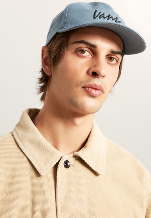 Casquette bleu clair avec le logo "Vans", portée avec une veste en velours côtelé beige dotée d'un grand col et de détails de bouton noir. Texture lisse.