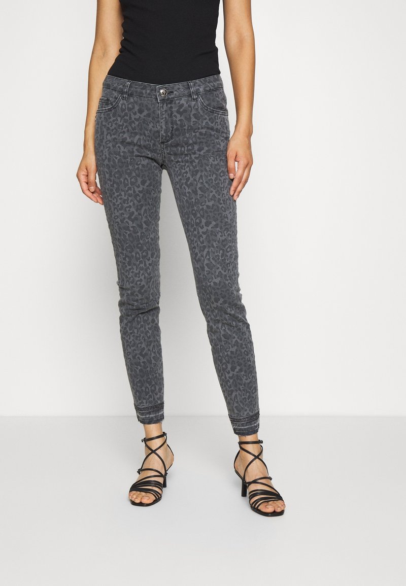Femme portant un jean slim gris à imprimé léopard et des sandales à talons noires à brides, debout devant un fond clair uni.