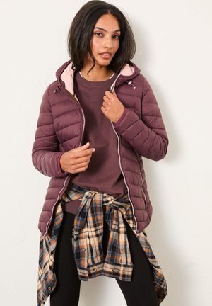 Bordeauxfarbene Pufferjacke mit einer rosa gefütterten Kapuze, Reißverschluss und gestepptem Textil, getragen über einem dunklen Oberteil und einem karierten Hemd, das um die Taille gebunden ist.