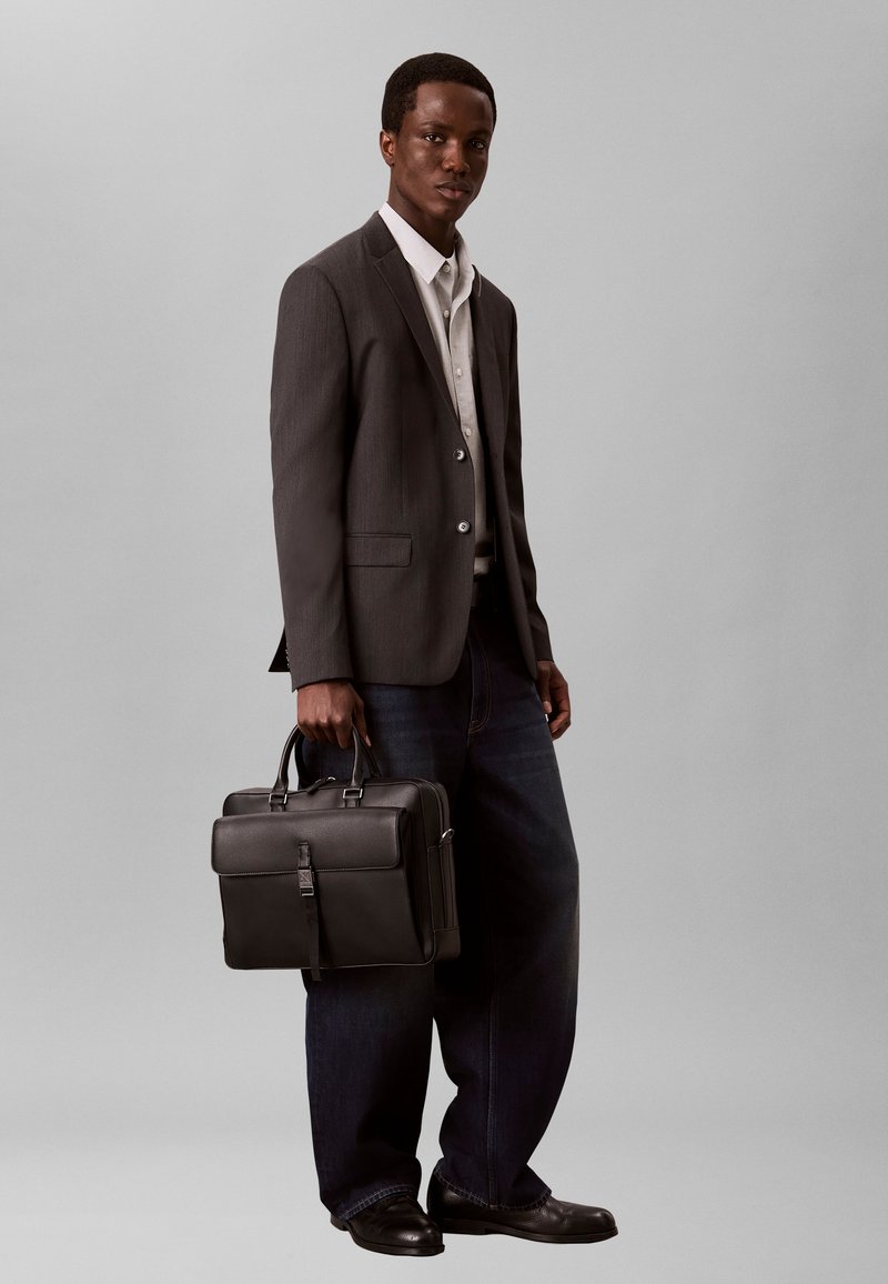 Homme portant un blazer sombre, une chemise blanche, un pantalon large foncé, des chaussures noires, tenant une mallette en cuir noir sur fond gris.