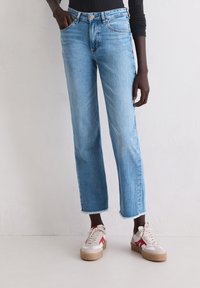 Personne portant un jean court bleu clair avec des ourlets effilochés et des baskets blanches avec des accents rouges, se tenant contre un mur blanc.