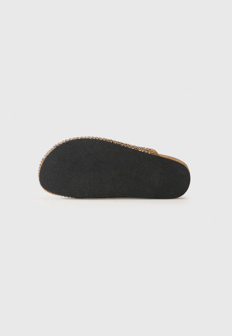 Sicht von unten auf einen braunen Slipper mit schwarzer strukturierter Gummisohle und Metallnieten entlang der Kante vor einem weißen Hintergrund.