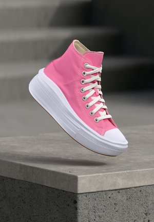 Zapatilla deportiva alta rosa con cordones blancos y suela blanca gruesa flotando sobre un borde de hormigón con un fondo de escaleras desenfocado.