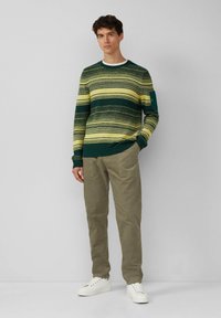 Man die tegen een effen grijze achtergrond staat, gekleed in een groen en geel gestreepte trui, kaki broek en witte sneakers.