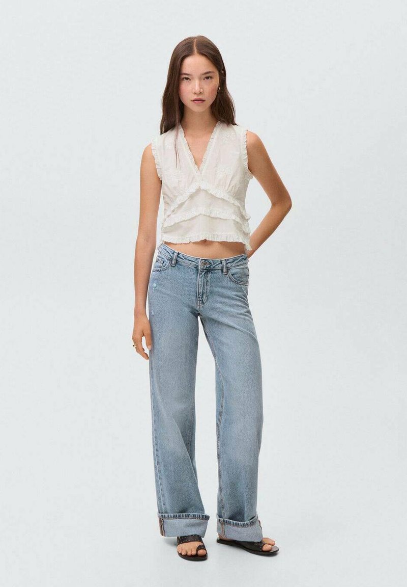 Un crop top blanc, à plusieurs couches, avec une texture froncée ; associé à un jean large bleu clair. Le mannequin se tient avec confiance, les mains sur les hanches.