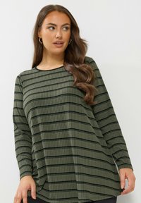 Nainen, jolla on pitkät, laineikkaat ruskeat hiukset, kullanväriset renkaat korvissa ja oliivinvihreä pitkähihaisvaate, jossa on mustia vaakaraitoja, katsoo sivulle.