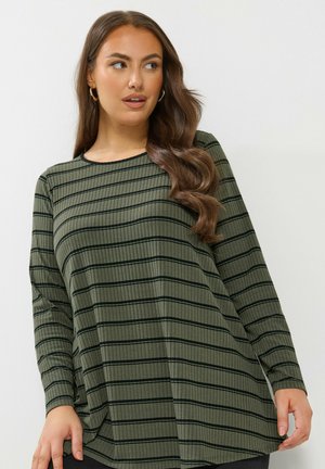 Vrouw met lang golvend bruin haar, draagt gouden oorbellen en een olijfgroen langel СолhngtОtop met zwarte horizontale strepen, kijkt opzij.