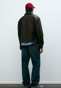 Mężczyzna w czerwonej czapce z daszkiem, ciemnobrązowej skórzanej kurtce, niebieskiej koszuli na guziki, niebieskich dżinsach i szarych butach, stojący twarzą do białej ściany.