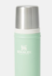 Mäta zelený Stanley termos s strieborným kovovým vrchom a bielym logom s nápisom "Stanley Since 1913" na plain bielom pozadí.