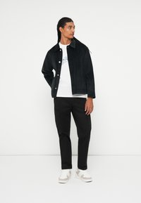 Sort fløjeljakke med knaplukning, hvid grafisk t-shirt, sorte cargobukser og lyse sneakers, stående mod en ensfarvet baggrund.
