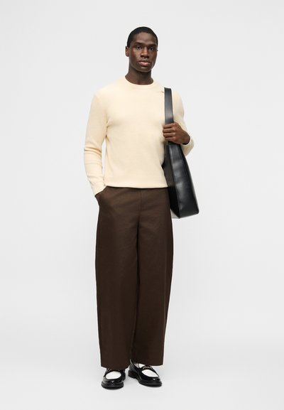 Homme portant un pull crème, un pantalon large marron, des mocassins noirs et blancs, portant un sac à bandoulière en cuir noir sur un fond clair.