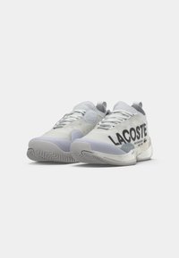 Άσπρα αθλητικά παπούτσια με κομψό σχέδιο, υφασμάτινη υφή, εξέχον λογότυπο "LACOSTE" σε μαύρο χρώμα και ελαφρώς γκρι σόλα από καουτσούκ.
