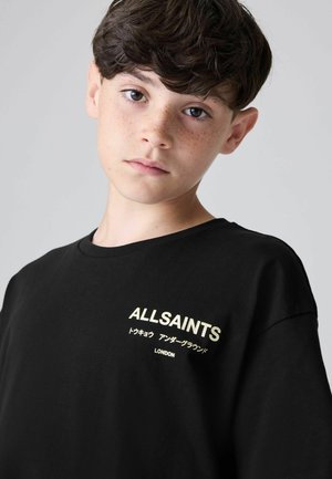Melna kokvilnas t-kreklā ar mazu zelta "ALLSAINTS" logo angļu un japāņu valodā, apaļš kakla izgriezums un īsas piedurknes.