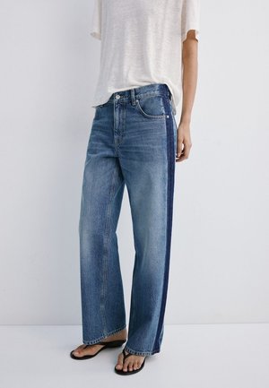 Person iført højtaljede, vidde blå jeans med en mørk sidestribe, kombineret med en løs hvid t-shirt og sorte flip-flops.