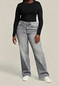 Personne portant un haut noir à manches longues, un jean large taille haute gris clair et des chaussures blanches sans lacets, debout sur un fond neutre.